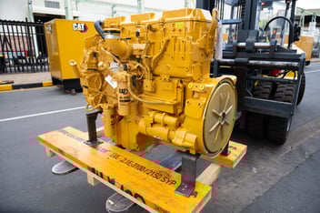 A bright yellow Caterpillar (CAT) industrial engine sits mounted on a wooden shipping pallet, supported by the forks of a forklift. The engine features complex components, including two visible CAT fuel filters and silver piping, all in pristine condition. The background shows an industrial yard with a paved surface and a yellow CAT generator unit.