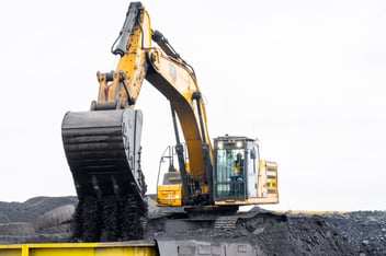 A medium shot of a yellow Caterpillar excavator atop a mound of coal, its bucket tilted to dump a load of dark material into a yellow truck bed. The machine is the focal point against a bright, overcast sky, highlighting the industrial scale of the operation.